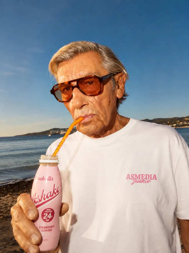 An older man in sunglasses on the beach in the white ASMEDIA Jaakie tee, drinking a strawberry milkshake