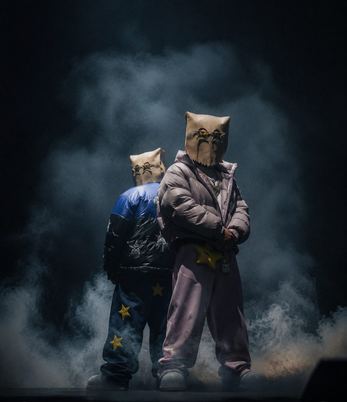 Two ASM paper-bag characters with star-shaped glasses standing in stage smoke, one in a lilac puffer jacket and pink trousers, the other in a blue and black star-print puffer and star-patched jeans